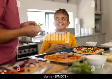 Der Mittelteil des weißen Großvaters mit dem süßen kleinen Enkel, der Pizza auf der Kücheninsel zubereitet Stockfoto