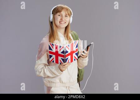 Lächelndes, trendiges Schulmädchen in beigefarbenem Trainingsanzug mit Rucksack, arbeitsbuch mit englischer Flagge und Kopfhörern mit Smartphone isoliert auf Grau. Stockfoto