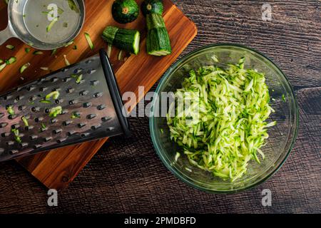 Geschredderte Zucchini in einer Rührschüssel aus Glas: Geriebene Zucchini, abgebildet mit einer Reibe und anderen Werkzeugen Stockfoto