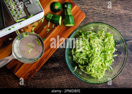 Geschredderte Zucchini in einer Rührschüssel aus Glas: Geriebene Zucchini, abgebildet mit Mandoline und anderen Werkzeugen Stockfoto