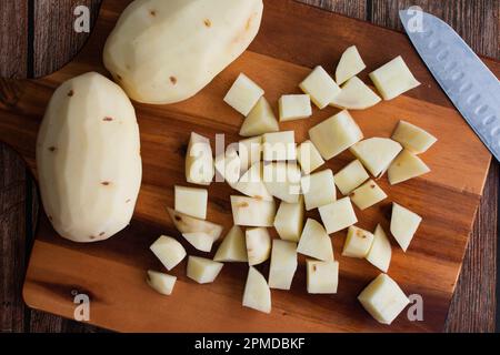 Geschälte und gewürfelte Russet-Kartoffeln auf einem Holzschneidebrett: Frische Kartoffeln, gehackt in kleine Würfel mit einem Küchenmesser und einem Schneidebrett Stockfoto