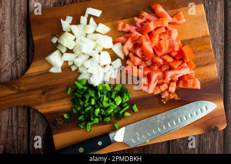 Gehackte Zwiebeln, Tomaten und grüne Chilipfeffer auf einem Holzschneidbrett: Mit einem Küchenmesser auf einem Holzschneidbrett vorbereitetes frisches Gemüse Stockfoto