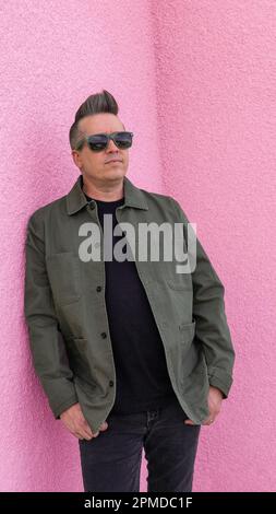 Ein Mann lehnt sich in einer Sonnenbrille gegen eine pinkfarbene Stuckwand und trägt schwarz mit einer grünen Jacke. Stockfoto