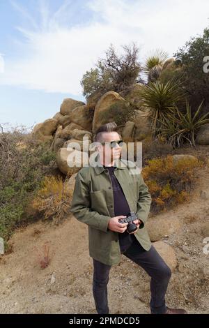Männlicher Tourist mit Sonnenbrille in Schwarz mit grüner Jacke und Kamera in Joshua Tree, Kalifornien. Stockfoto