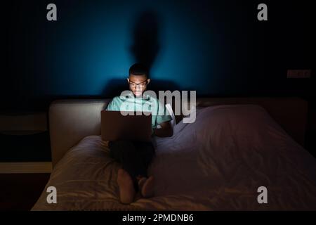Ein junger Schwarzer arbeitet mit einem Laptop, der nachts im Bett liegt. Er sieht aufmerksam auf den Bildschirm. Stockfoto