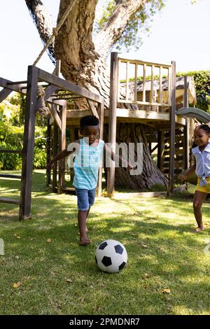 Fröhliche afroamerikanische Geschwister, die auf dem grasbedeckten Feld im Park Fußball spielen Stockfoto