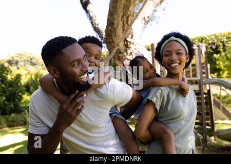 Fröhliche afroamerikanische Eltern, die ihren Sohn und ihre Tochter auf dem Spielplatz mit dem Huckepack betreuen Stockfoto