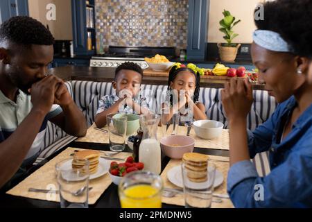 afroamerikanische Eltern und Kinder, die morgens vor dem Frühstück am Esstisch das Tischgebet sprechen Stockfoto