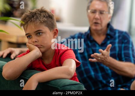 Kaukasischer trauriger Junge mit Hand am Kinn, der wegschaut, während er mit Großvater auf dem Sofa sitzt Stockfoto