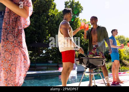 Fröhliche, vielfältige Gruppe von Freunden, die eine Poolparty veranstalten, zusammen im Garten grillen Stockfoto