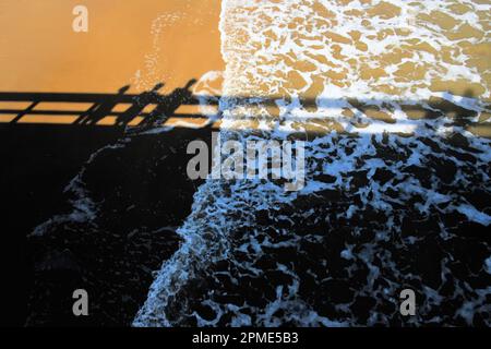 Ein Foto von Sand und Wasser mit einer Reflexion von drei Personen, die auf einem Pier in South Padre Island, Texas, USA, stehen. Stockfoto