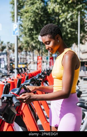 Trendige afrikanische Frau, die ihr Telefon benutzt, um ein öffentliches System des Fahrradverleihs zu nutzen Stockfoto
