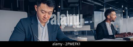 Fokussierter asiatischer Geschäftsmann, der Kaffee trinkt, während er im modernen Büro mit Dokumenten arbeitet Stockfoto
