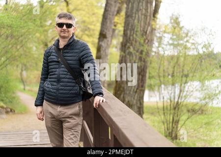 Ein Mann mittleren Alters mit Sonnenbrille und einer dünnen Daunenjacke auf einer hölzernen Fußgängerbrücke in der Nähe des Sees im Frühling Stockfoto