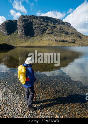 Wanderer mit Rucksack am Ufer des Loch na Creitheach mit den Felsen von Sgurr Hain in The Black Cuillins Beyond, Camasunary, Skye, Großbritannien Stockfoto