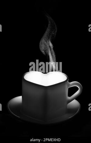 Ein weißer herzförmiger Becher mit einem warmen Getränk und ausgefallenem lockigem Dampf. Eine dampfende Kaffeetasse auf schwarzem Hintergrund. Morgenkaffee. Kaffee für Stockfoto