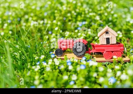 Miniaturbewegung: Spielzeugtraktor, der ein winziges Haus auf einer Wiese mit frischem grünen Gras und bunten Blumen trägt. Heller Hintergrund für die Freude, Fre zu starten Stockfoto