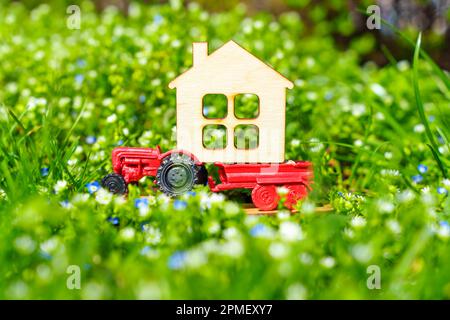 Miniaturtraktor mit einem Flachhausmodell auf einem Blumenbeet aus üppigem grünen Gras und bunten Frühlingsblumen. Frühling als Saison für einen neuen Stockfoto