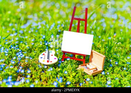 Winzige Leinwand auf einem kleinen Standfuß, Miniaturpalette und Holzkiste mit Farben und Pinseln in einem üppigen blühenden Feld. Kunstbedarf und Ausrüstungskonzept. Stockfoto