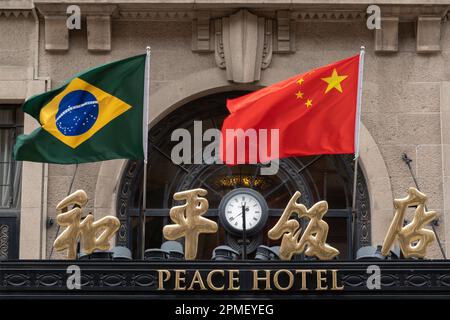 SHANGHAI, CHINA - 13. APRIL 2023 - Chinesische und brasilianische Nationalflaggen flattern im Wind über dem Haupteingang des Peace Hotels an der Nanjing Road Stockfoto