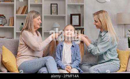 Eine ältere Frau, eine Großmutter und eine junge Mutter machen Zöpfe für eine kleine blonde Enkelin auf dem Sofa im Wohnzimmer. Zeit mit der Familie verbracht Stockfoto