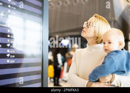 Mutter reist mit Kind, hält seinen kleinen Jungen am Flughafenterminal, überprüft Flugplan, wartet auf das Einsteigen in ein Flugzeug. Das Konzept „Reisen mit Kindern“ Stockfoto