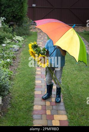 Ein unbekannter Junge mit einem Strauß Sonnenblumen versteckt sich hinter einem bunten Regenschirm. Regnerisch bewölktes Wetter. Ein Blumengeschenk für mom Stockfoto