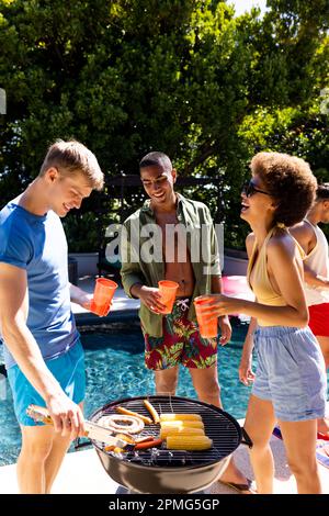 Fröhliche, vielfältige Gruppe von Freunden, die eine Poolparty veranstalten, zusammen im Garten grillen Stockfoto