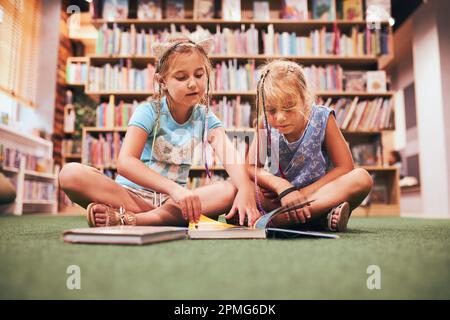 Zwei Schulmädchen lesen Bücher in der Schulbibliothek. Grundschüler lernen aus Büchern. Schüler machen Hausaufgaben. Kinder, die Spaß mit Büchern haben Stockfoto