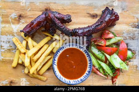 Gegrillte geräucherte Schweinerippchen mit pommes Frites, Sauce und Salat auf Holzbrett auf schwarzem Hintergrund, Draufsicht Stockfoto