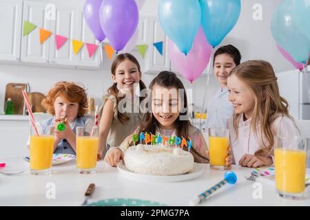Ein fröhliches Mädchen, das sich Geburtstagskuchen in der Nähe einer glücklichen Gruppe von Freunden anschaut, während der Party zu Hause, Stock Image Stockfoto