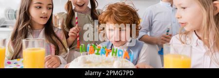 Rothaariger Junge bläst Kerzen auf Geburtstagskuchen in der Nähe von Freunden während der Party zu Hause, Banner, Stock Image Stockfoto