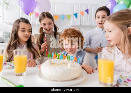 Rothaariger Junge bläst Kerzen auf Geburtstagskuchen in der Nähe von Freunden während der Party zu Hause, Stock Image Stockfoto