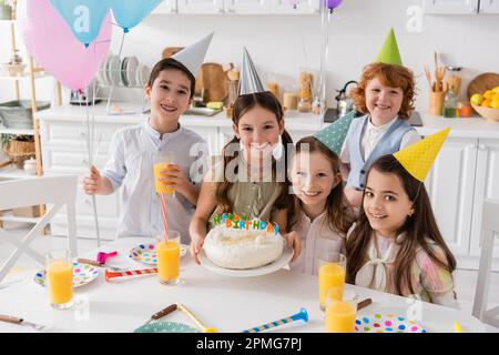 Eine Gruppe fröhlicher Kinder in Partykappen, die Spaß beim Geburtstag zu Hause haben, Stock Image Stockfoto