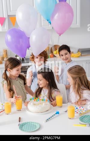 Fröhliche Kinder mit Luftballons, die ein Geburtstagskind in der Nähe von Kuchen und Partyhörnern auf dem Tisch anschauen, Stockbild Stockfoto