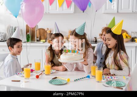 Glückliches Mädchen, das Geburtstagskuchen mit Kerzen in der Nähe von fröhlichen Freunden hält, während der Feier zu Hause, Stock Image Stockfoto