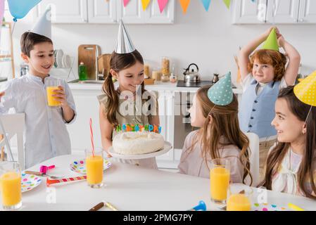 Glückliches Mädchen, das Geburtstagskuchen mit Kerzen in der Nähe von fröhlichen Freunden hält, während der Party zu Hause, Stock Image Stockfoto