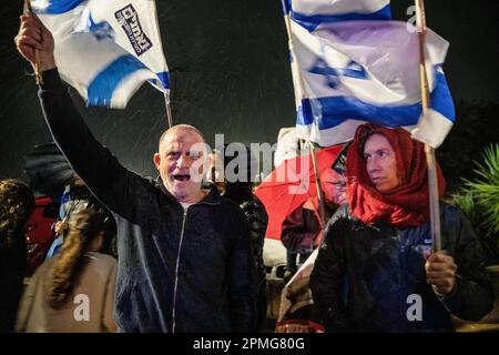 Hadera, Israel. 12. April 2023. Reformfeindliche Demonstranten schwenken israelische Flaggen und halten während der Demonstration Schilder im Regen. Demonstranten für und gegen die Justizreform in Hadera demonstrieren vor einer Mimouna-Zeremonie unter Teilnahme von Ministerpräsident Benjamin Netanjahu, dass es Konfrontationen zwischen reformfeindlichen Demonstranten und reformfreundlichen rechtsgerichteten Aktivisten gab. Kredit: SOPA Images Limited/Alamy Live News Stockfoto