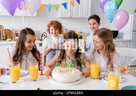 Glückliches Mädchen, das sich ihren Geburtstagskuchen mit Kerzen in der Nähe von Freunden ansieht, während der Feier zu Hause, Stock Image Stockfoto