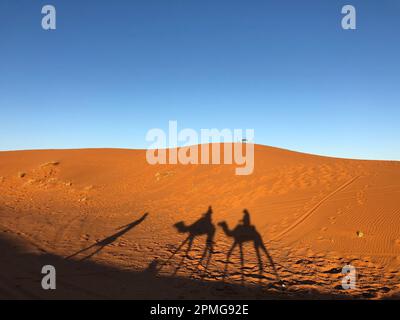 Zwei Personen reiten auf Kamelen in einer Wüstenlandschaft, mit einer großen Sanddüne im Hintergrund Stockfoto