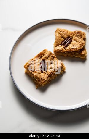 Zwei Quadrate Blondie Kekse mit Pekannüssen auf weißem Teller. Weiße Schokolade, süß und lecker, kaute Keksriegel. Schließen Stockfoto