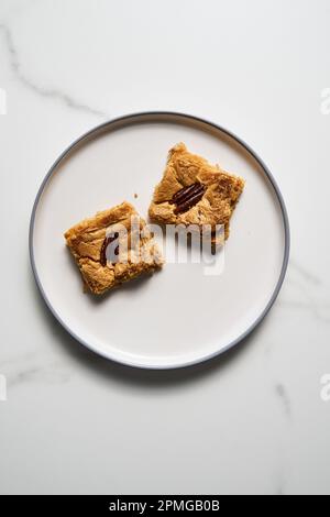 Zwei Quadrate Blondie Kekse mit Pekannüssen auf weißem Teller. Weiße Schokolade, süß und lecker, kaute Keksriegel. Draufsicht, Kopierbereich Stockfoto