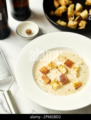 Biersuppe mit Croutons und Muskatnuss. Daneben stehen Flaschen Bier, Croutons und eine Reibe. Weißer Hintergrund Stockfoto