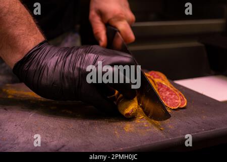 Nicht wiedererkennbare Metzgerhände, die Wurst auf einem Schneidebrett schneiden, selektiver Fokus auf dem schwarzen Handschuh. Stockfoto