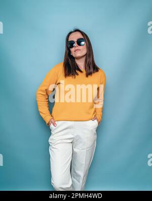 Braune Frau mit dunkler Brille, gelber Pullover steht Hände in Taschen auf blauem Hintergrund. Stockfoto