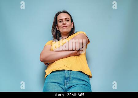 Eine junge Frau aus dem unteren Winkel kreuzte die Arme auf blauem Hintergrund Stockfoto