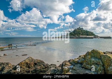 St. Michaels Mount vor der Küste von Cornwall in der Nähe von Marazion an einem klaren sonnigen Tag im April. Felsen im Vordergrund und Menschen, die über das Meer laufen. Stockfoto