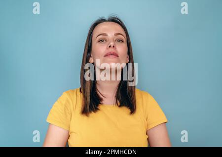 Die junge Frau schaut mit erhobenem Kopf in die Kamera. Trägt ein gelbes T-Shirt auf blauem Hintergrund Stockfoto