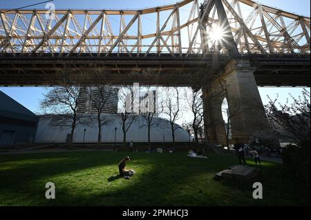 New York, USA. 12. April 2023. Licht gefiltert durch die 59. Street Bridge Struktur beleuchtet eine Frau, die in einem kleinen Park auf Roosevelt Island, New York, NY, am 12. April 2023 sitzt. Die Temperaturen im Sommer werden in New York City voraussichtlich 90 Grad erreichen und damit die bisherigen Rekorde für den Monat April brechen. (Foto: Anthony Behar/Sipa USA) Guthaben: SIPA USA/Alamy Live News Stockfoto