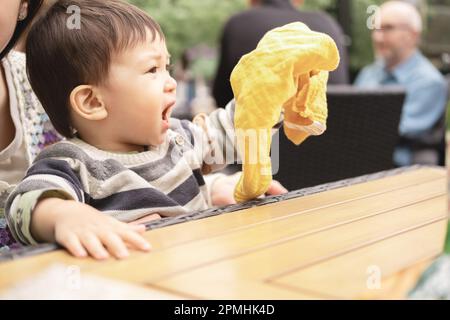 Ein männlicher Säugling, der auf den Beinen ihrer mutter sitzt, schreit und schüttelt mit der Hand ein gelbes Tuch. Multikulturelle und multirassische Familie, die ein Bier trinkt Stockfoto